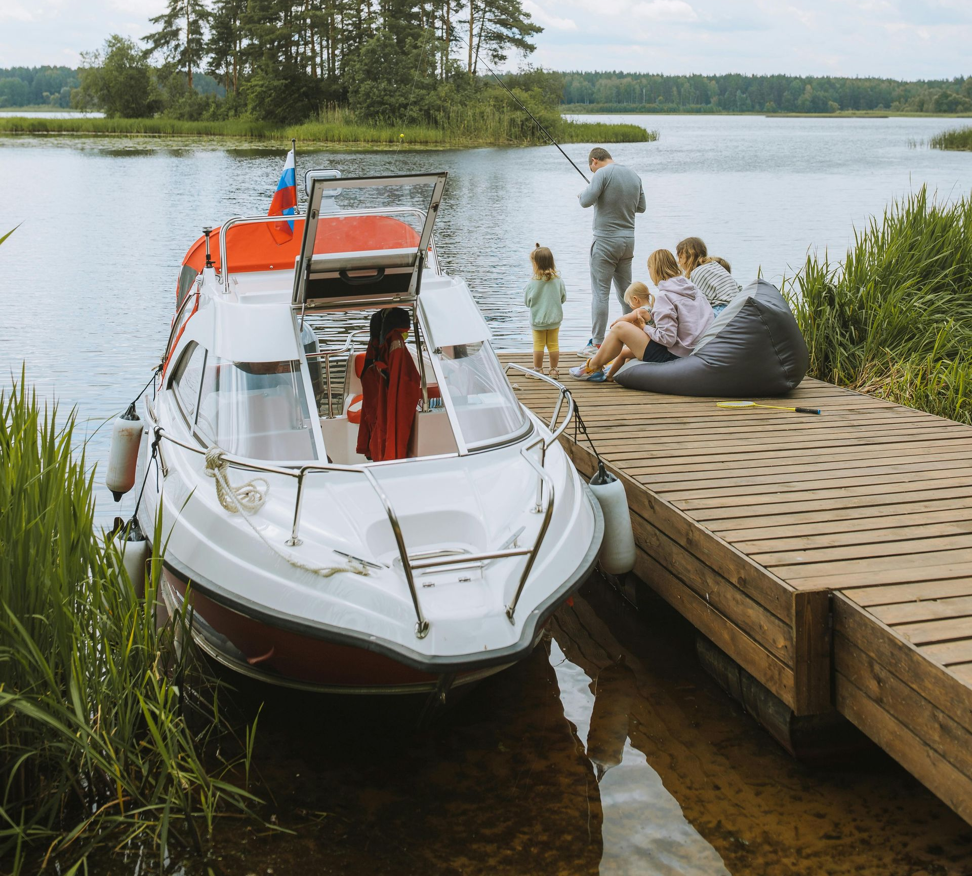 Boat docked at wooden pier with a family enjoying the lake. Green reeds and trees in the background.