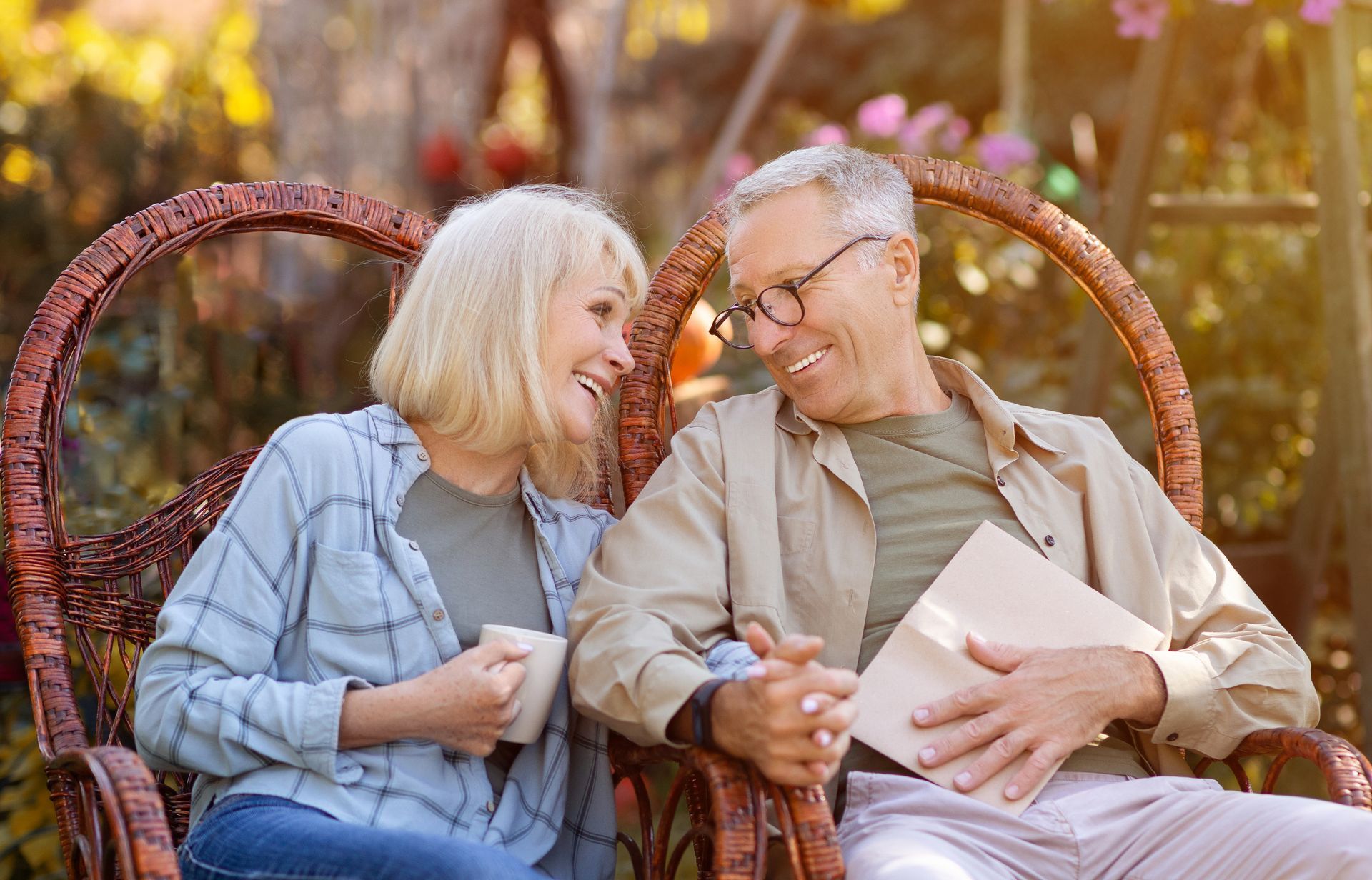 Couple sitting in wicker chairs, laughing. Woman holds mug; man holds a book outdoors.