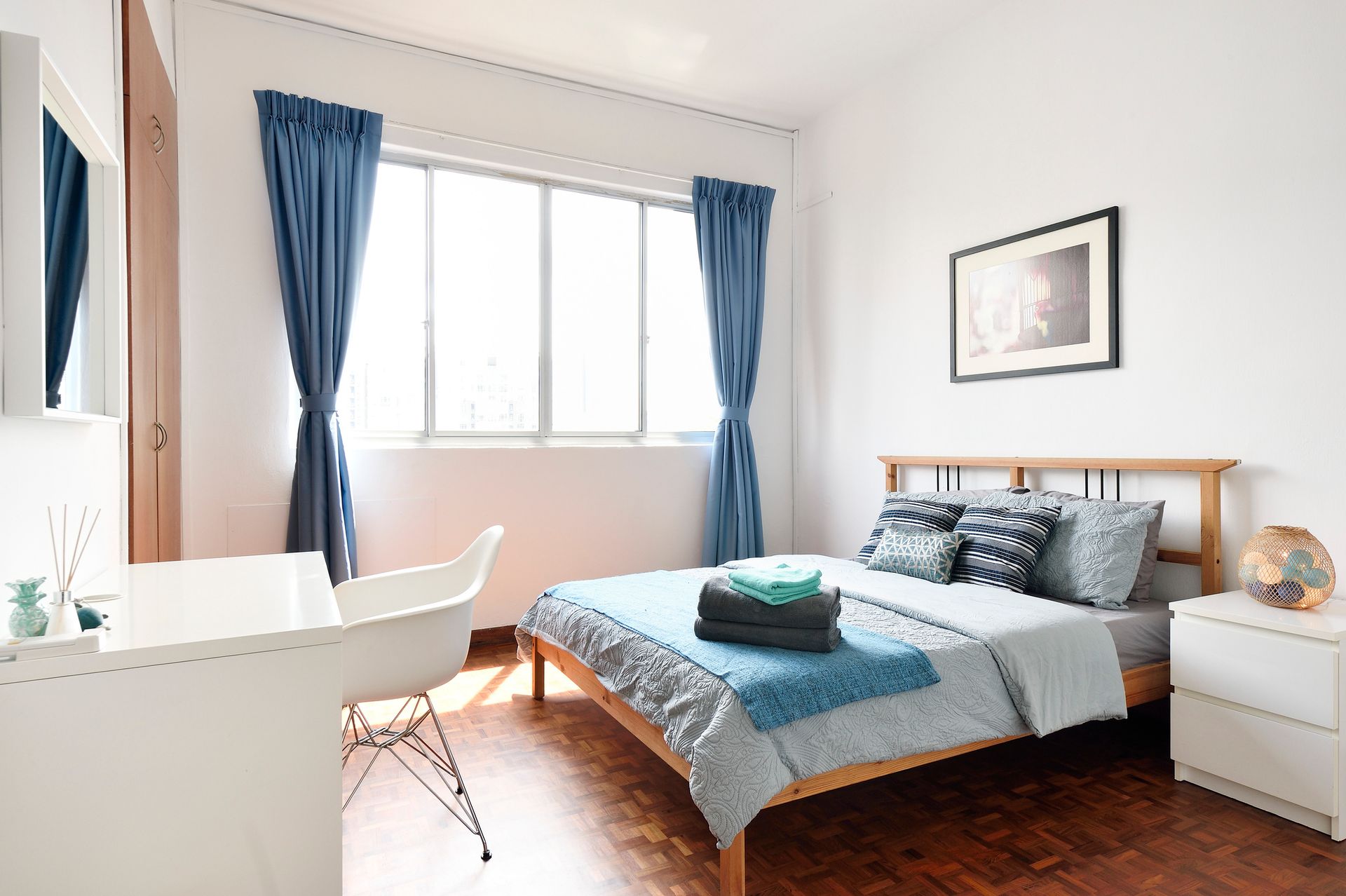 Bedroom with bed, desk, and blue curtains. Bright, wooden floors and white walls.