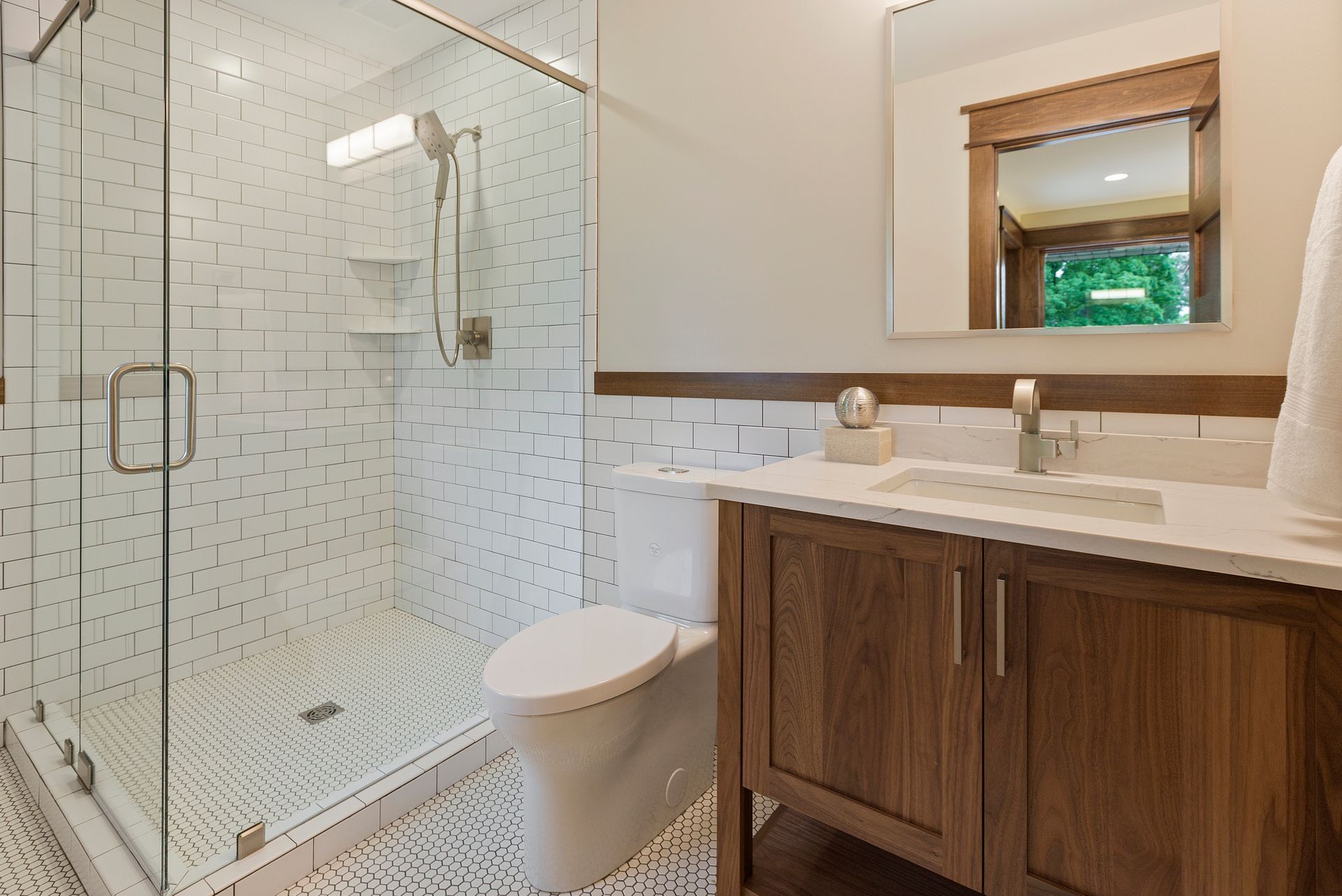Modern bathroom with white and brown tiling, glass shower, wooden vanity, and toilet.