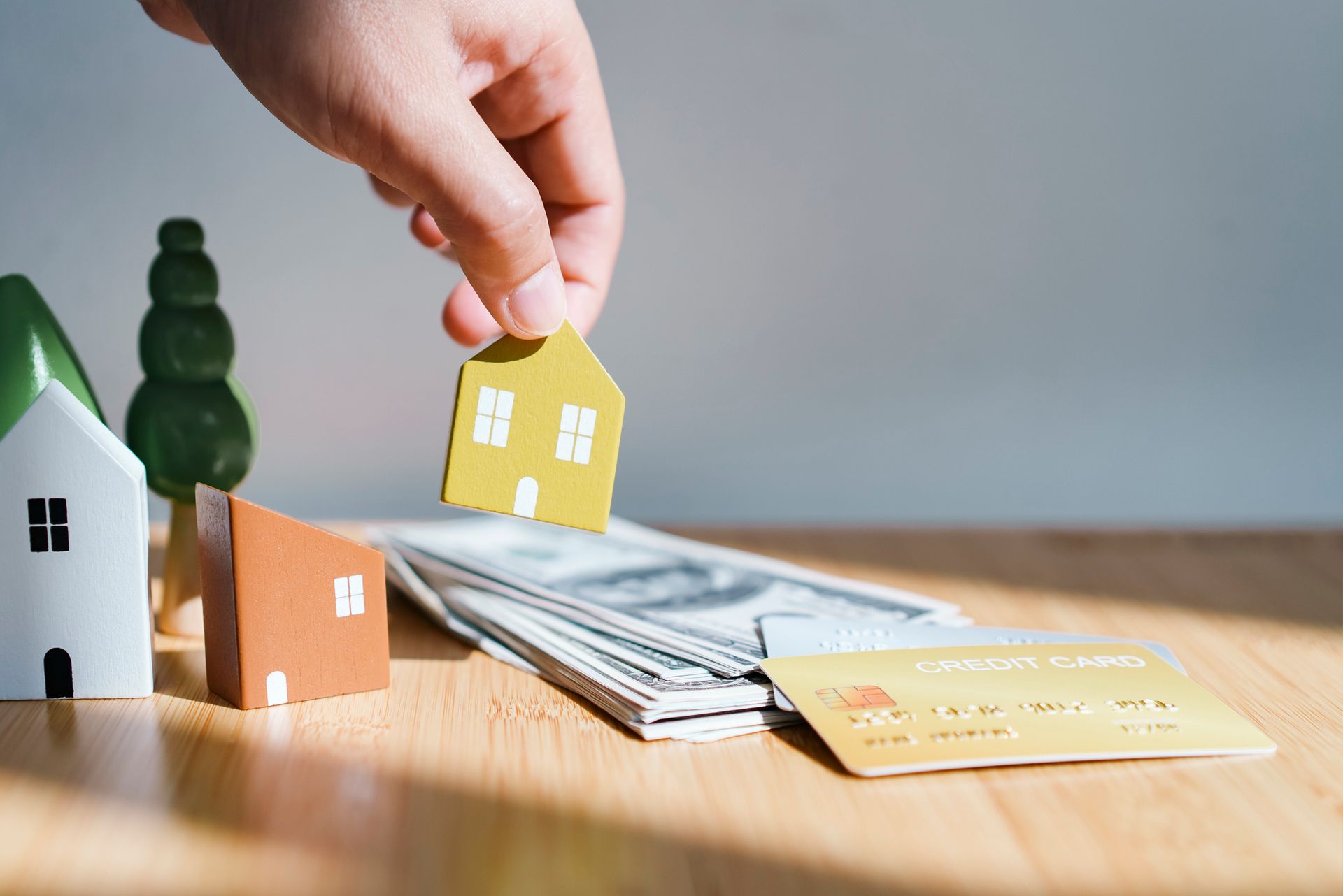 Hand holding a yellow house model over cash and credit card, with other house models on wood table.