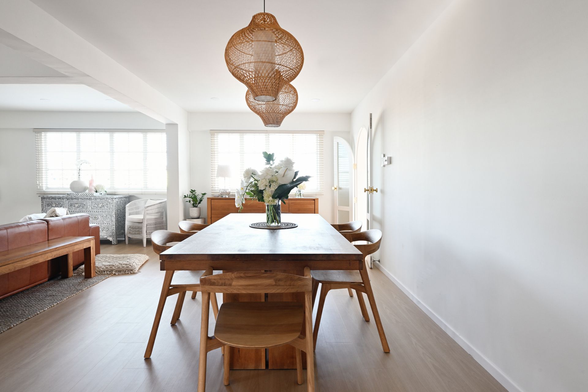 Wooden dining table set in a bright room, under two woven pendant lights.