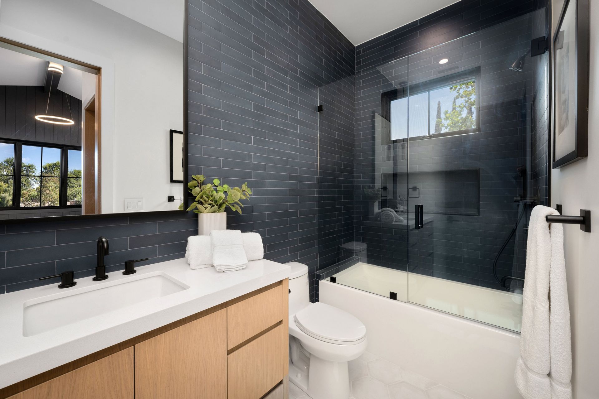 Modern bathroom with dark blue tiled wall, glass shower, wood vanity, and white fixtures.