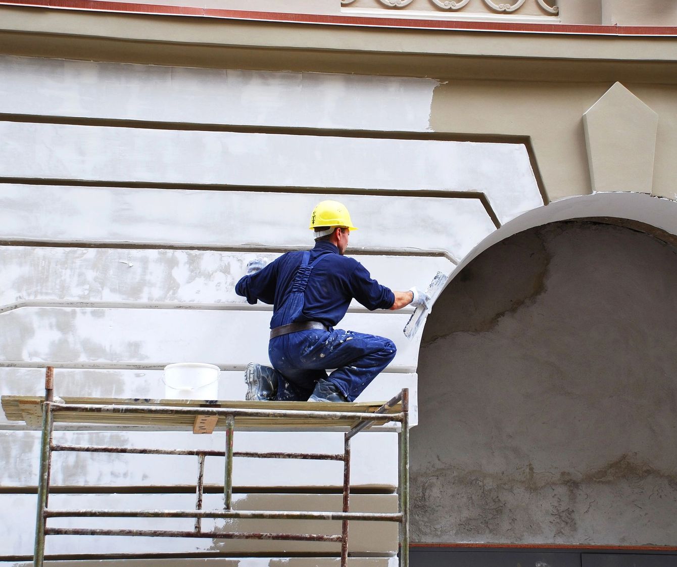 Un operaio con un elmetto giallo dipinge la facciata di un edificio da un'impalcatura.