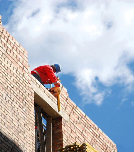 Un operaio con giacca rossa e casco blu si sporge da un edificio di mattoni sotto un cielo azzurro.