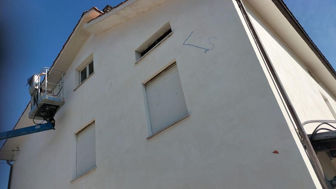 Edificio in stucco bianco con persiane chiuse, visto dal basso contro un cielo azzurro e un ascensore con balcone.