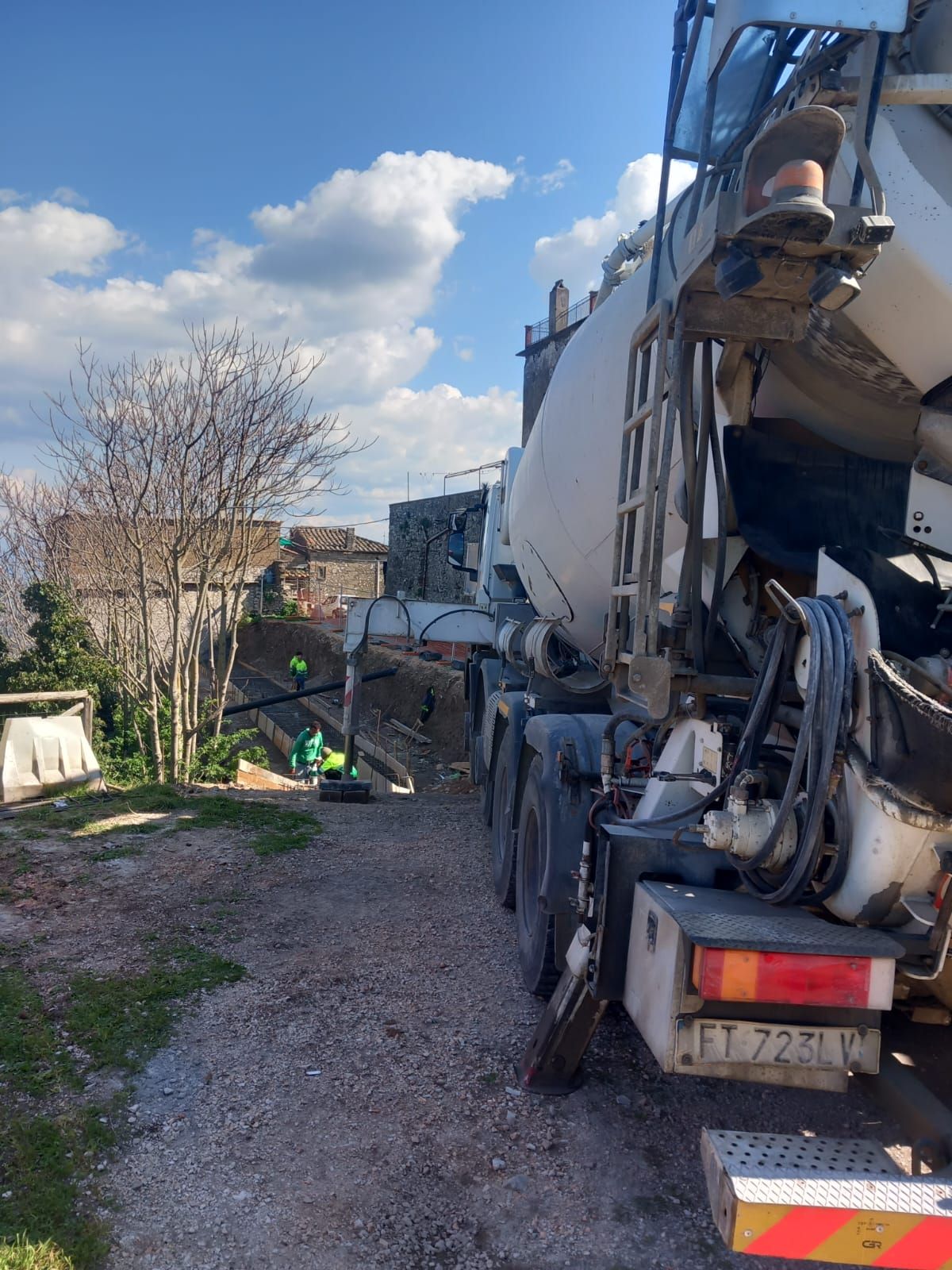 Autobetoniera in un deposito di ghiaia accanto a un edificio, con tubi e attrezzature sotto un cielo nuvoloso.