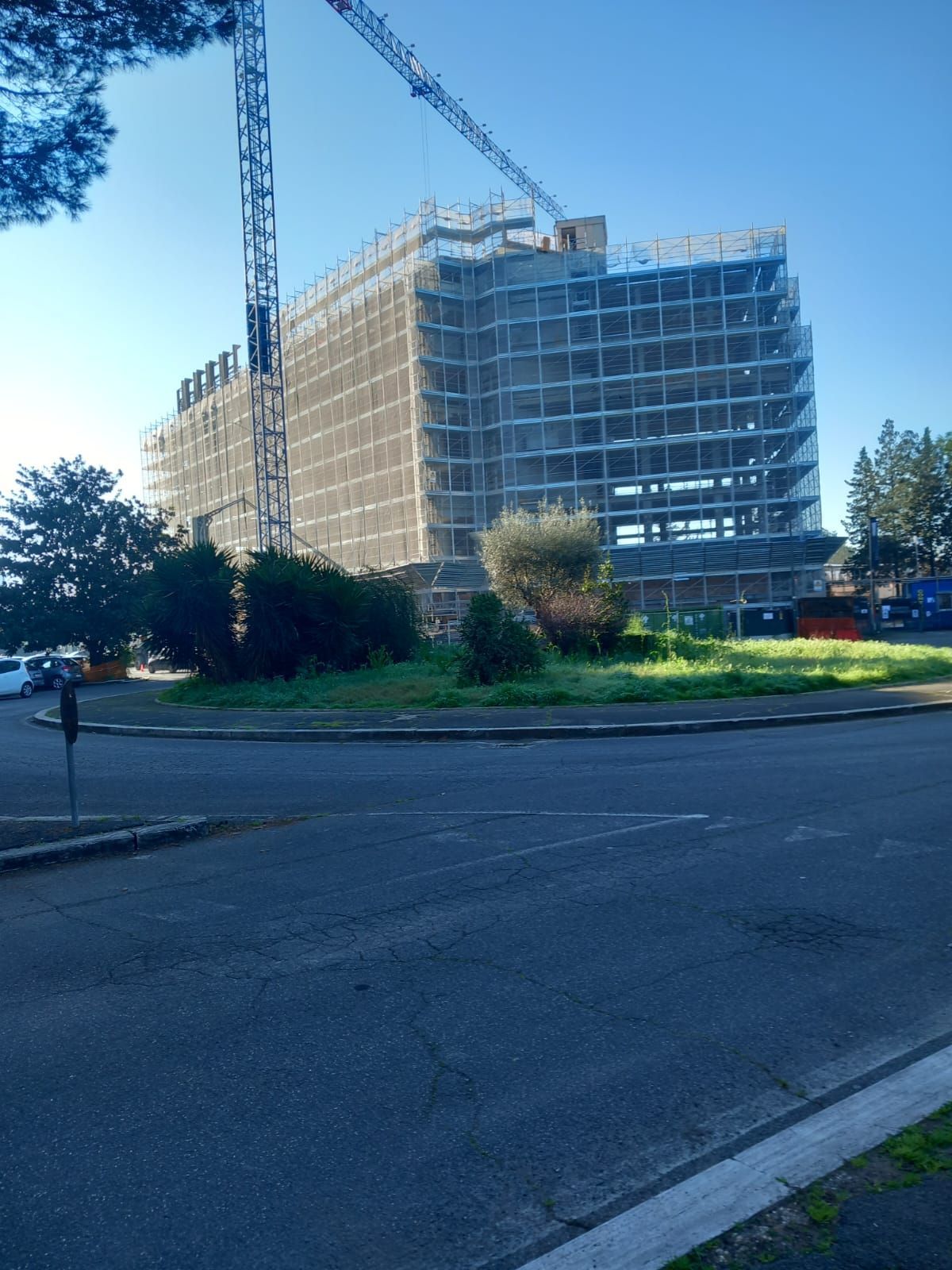 Edificio a più piani in costruzione, con impalcature, a lato di una strada, con alberi e cielo azzurro sullo sfondo.
