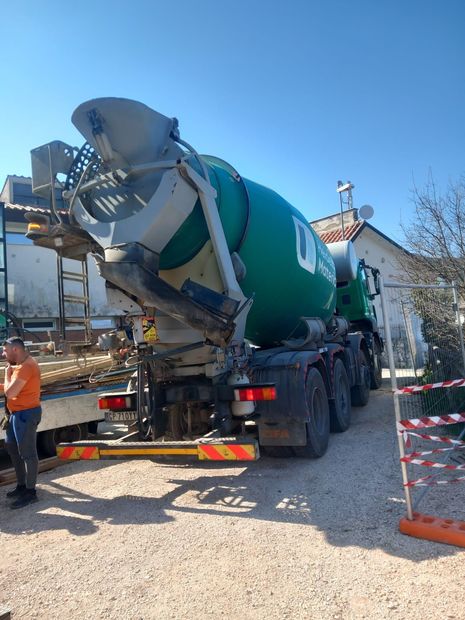 Un camion betoniera verde in un cantiere, con un operaio nelle vicinanze e barriere di sicurezza rosse e bianche.