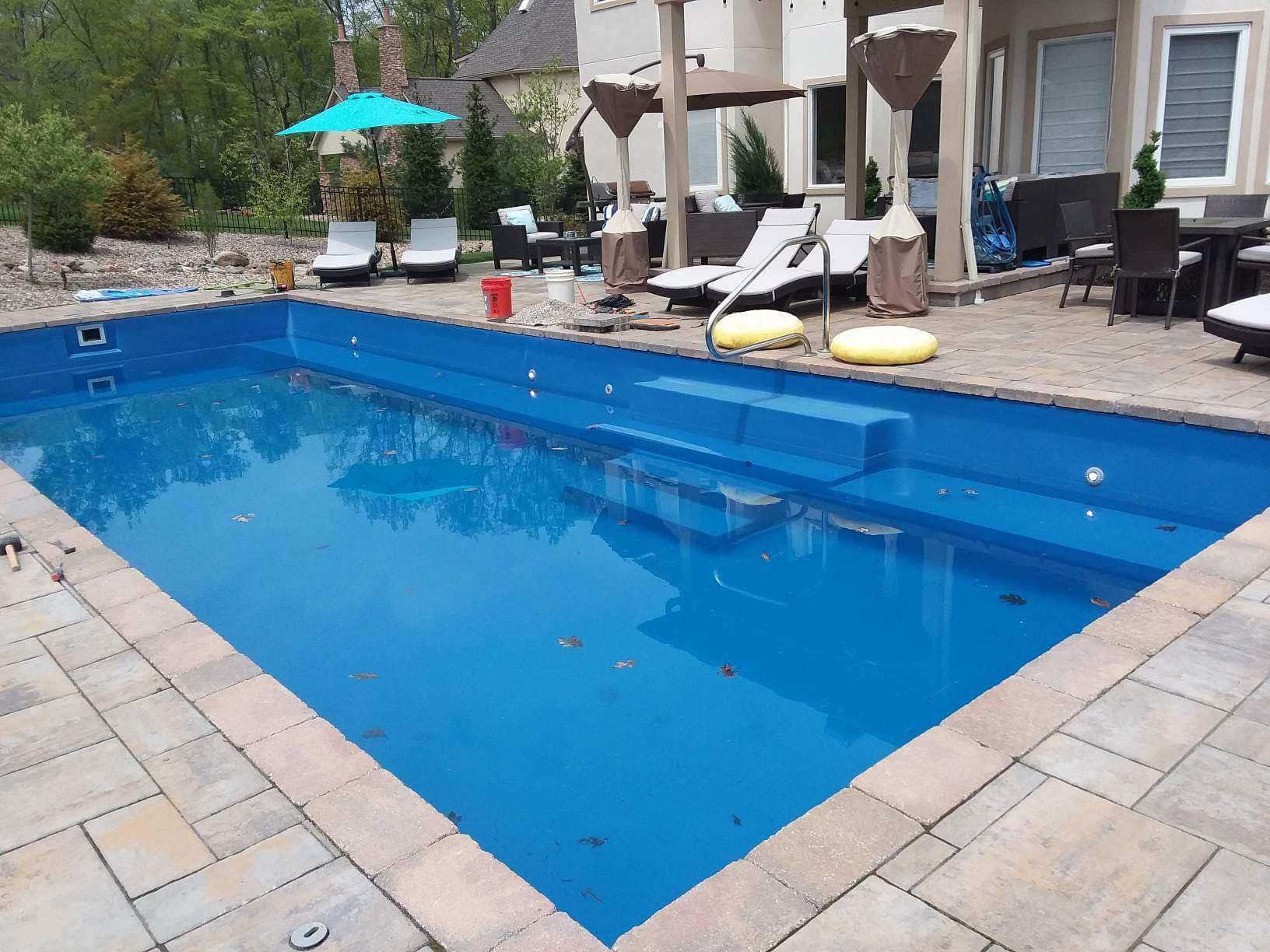 A rectangular blue pool with stairs, surrounded by paving stones and lounge chairs; sunny outdoor setting.