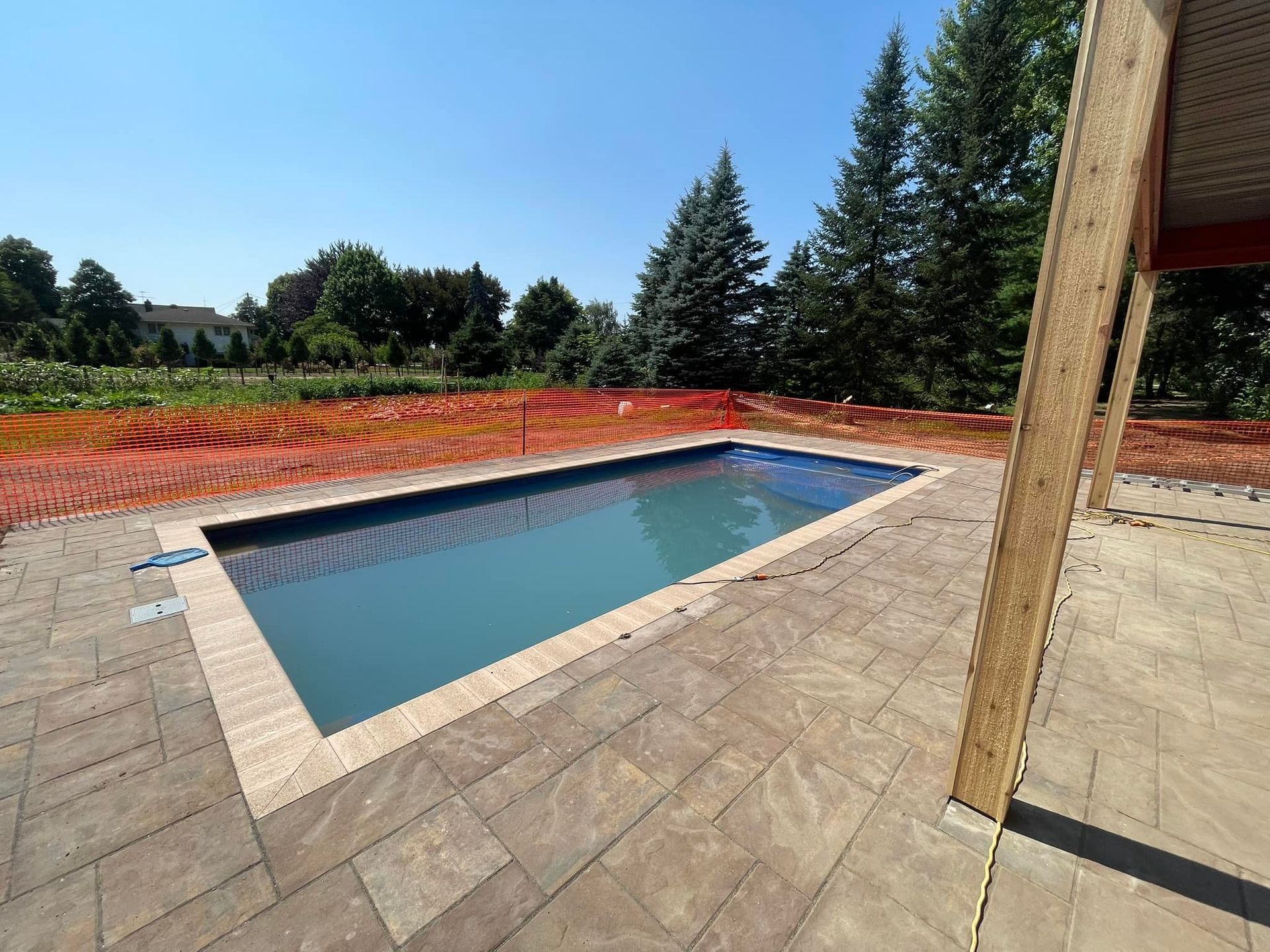 Rectangular pool surrounded by stone patio; blue water, trees, blue sky.