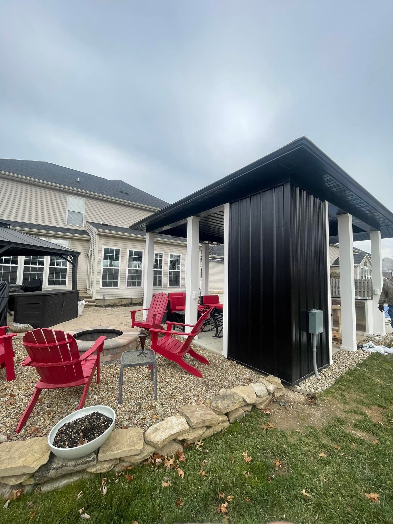 Gazebo with black corrugated siding, red chairs, and fire pit on a patio next to a house.