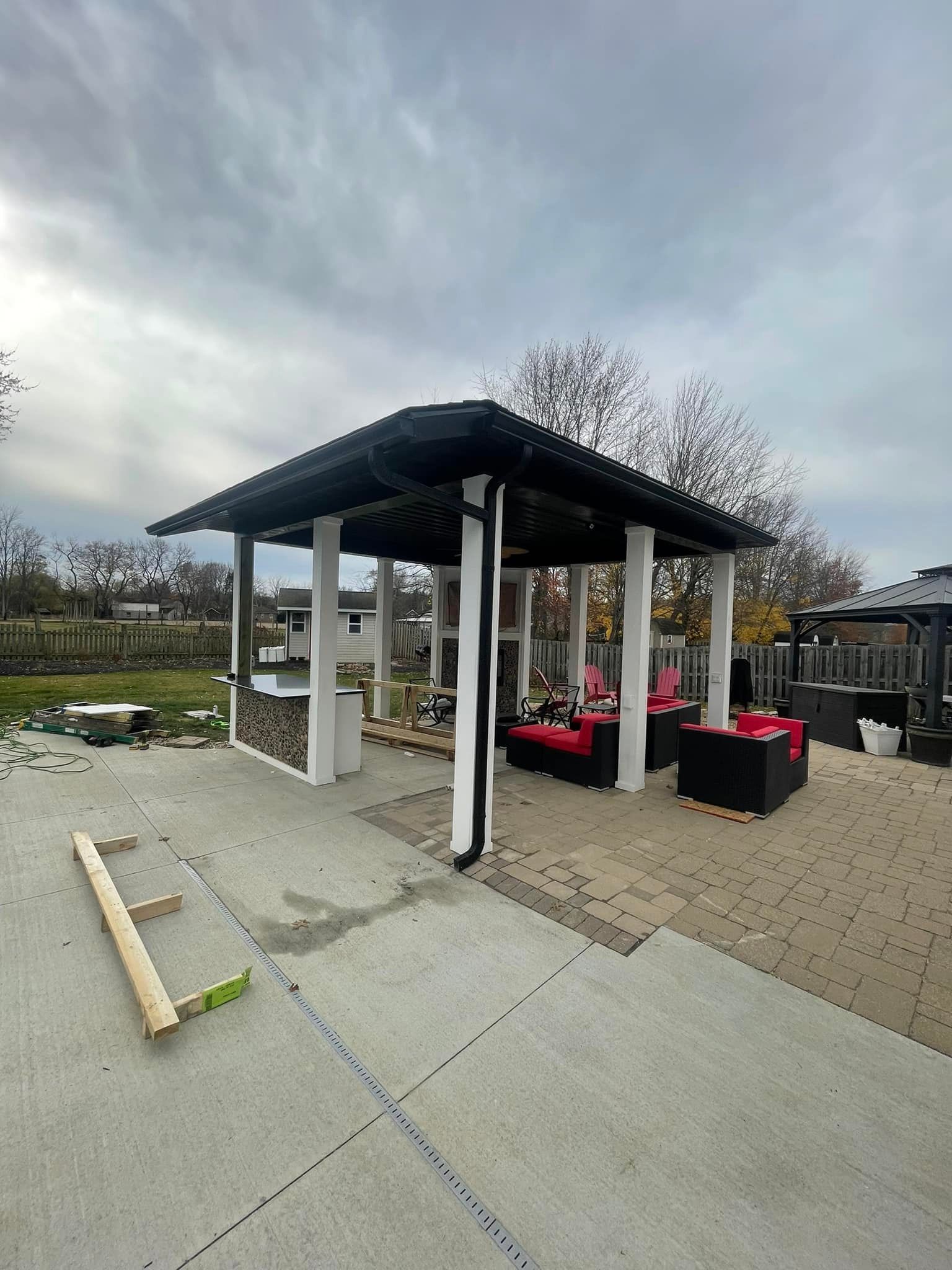 Outdoor pavilion with black roof, white columns, and red seating on a patio with overcast sky.