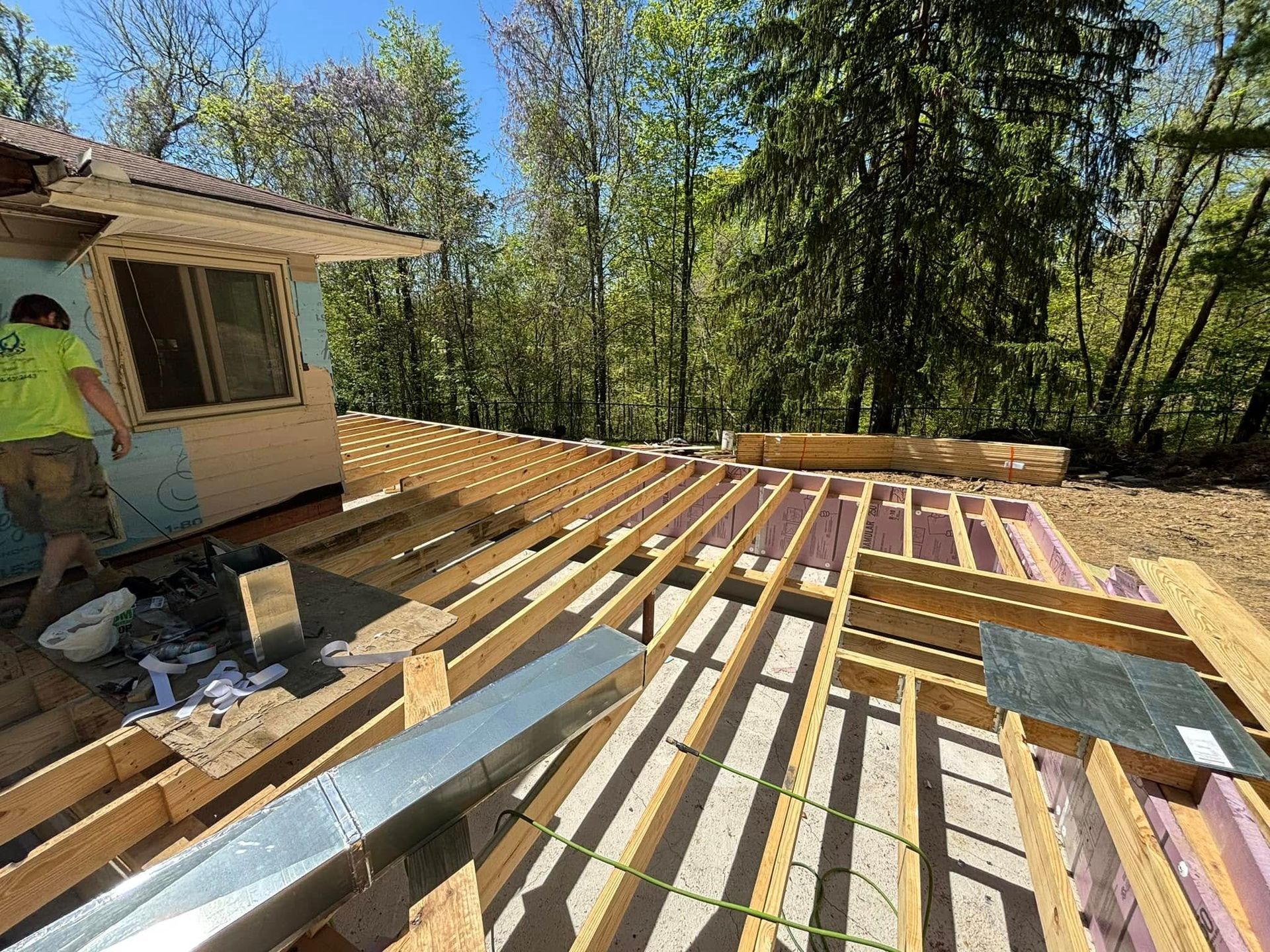 Construction site with wooden beams forming a floor, a worker near a window, and trees in the background on a sunny day.