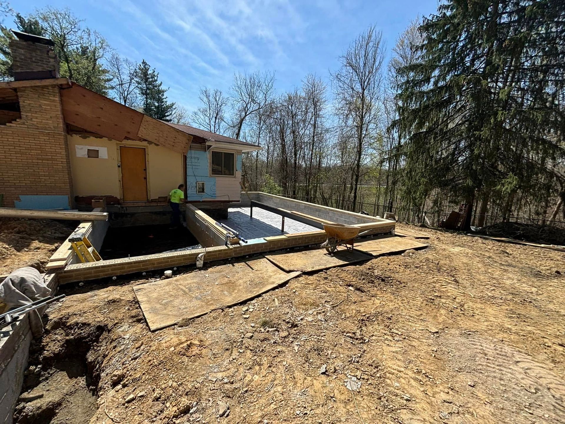 Construction site: foundation being built next to a beige house with a chimney. Bright, sunny day.