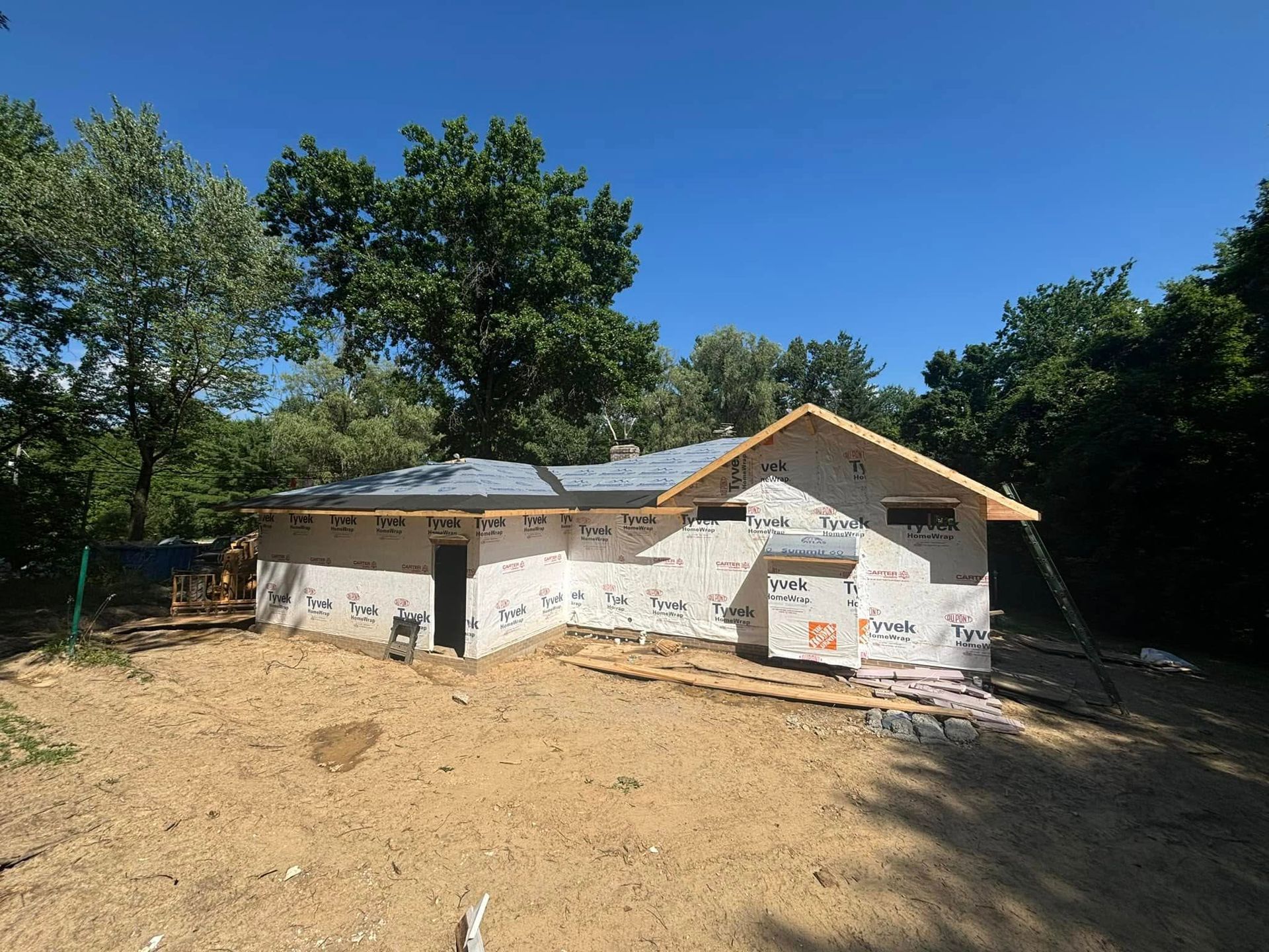 Construction of a one-story house, walls wrapped in white material, surrounded by trees and dirt, under a blue sky.