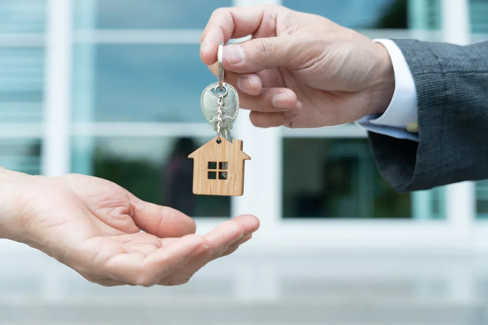 Hand passing house keys with a wooden house-shaped keychain in front of a building.