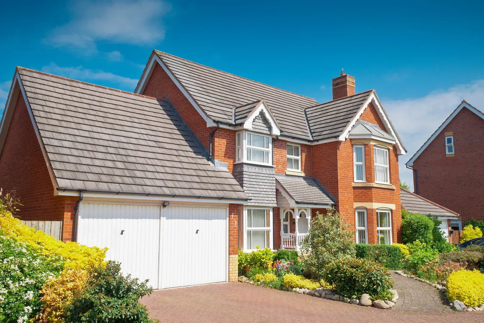 Brick house with a gray roof, white garage doors, and a well-maintained garden under a blue sky.