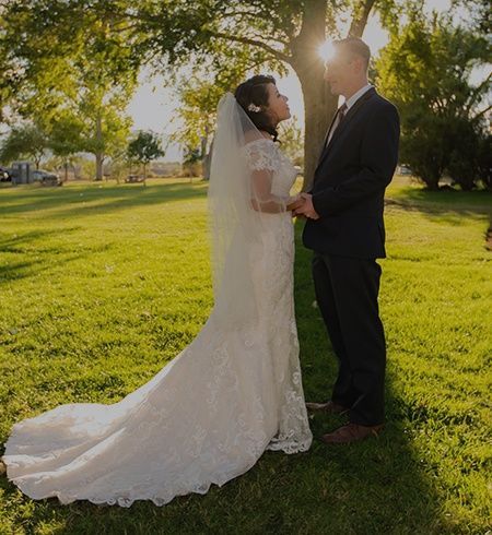 A bride and groom are standing under a tree in a park holding hands.