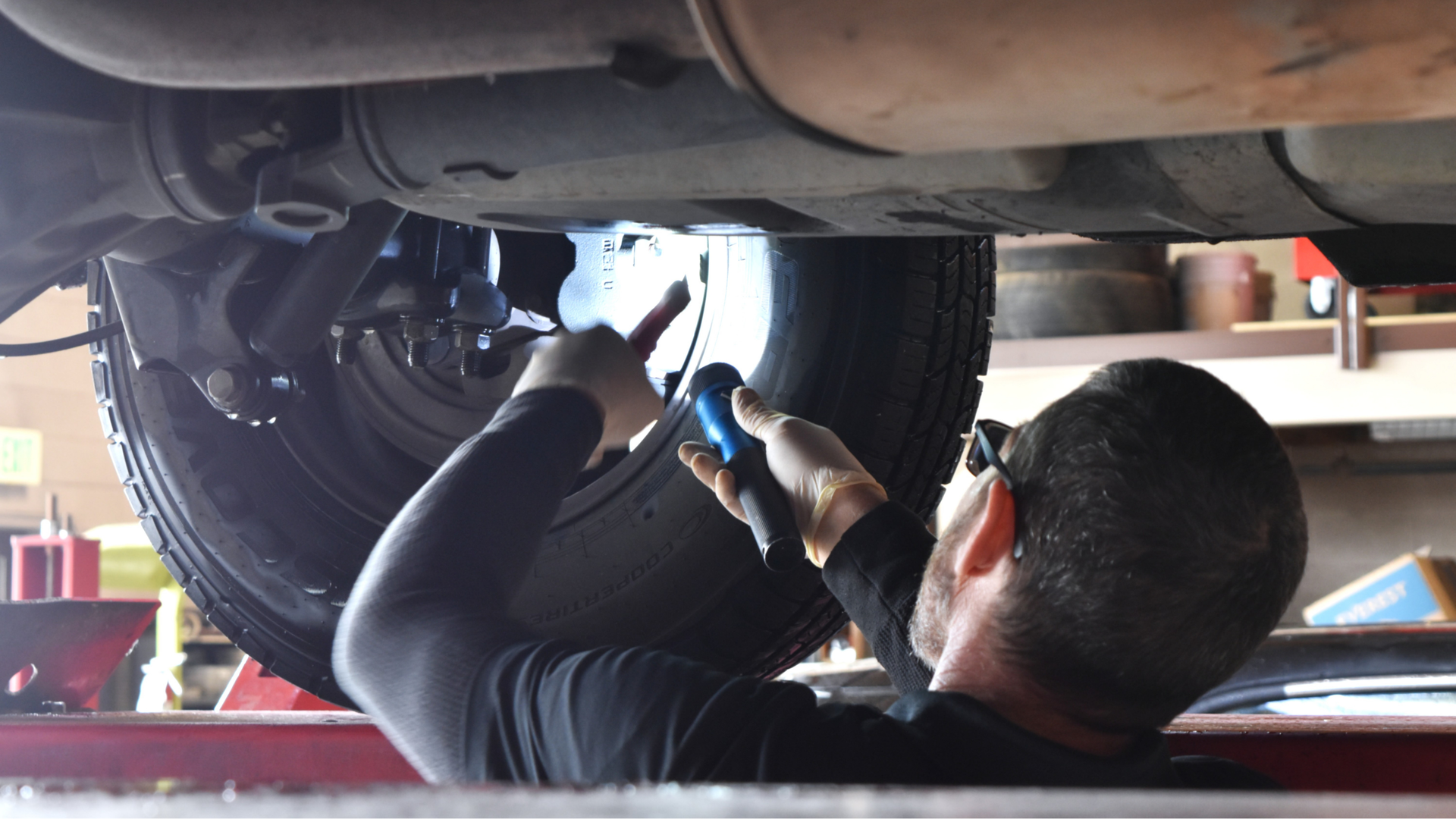 Precision Tire employee using a flashlight to inspect car brakes under the vehicle, ensuring safety