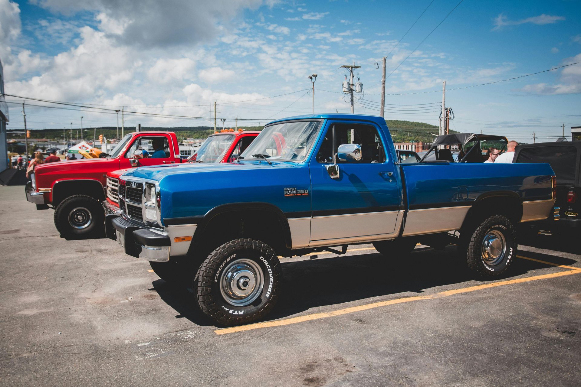 Blue and beige lifted pickup truck parked outdoors with red trucks behind it under a partly cloudy sky