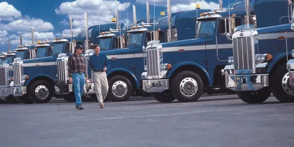 Two men are standing in front of a row of blue semi trucks.