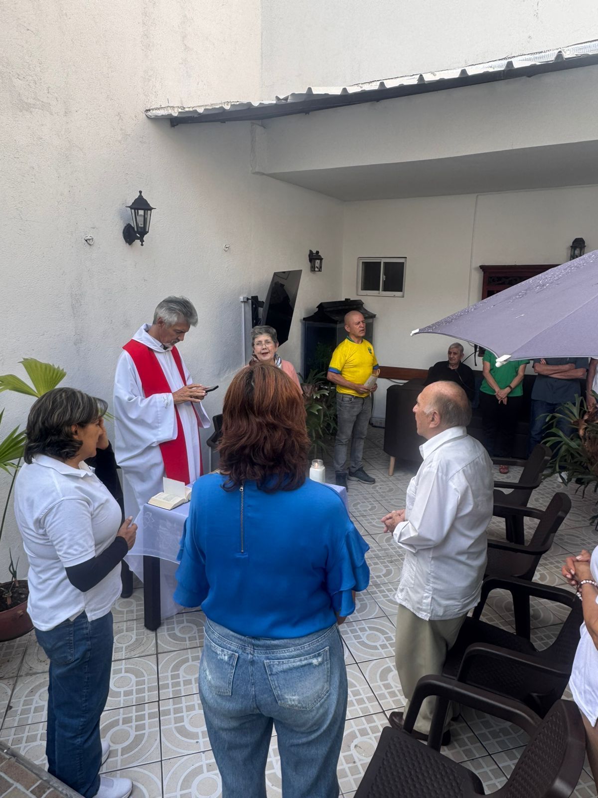 Un grupo de personas se reunió para una ceremonia religiosa al aire libre. Un sacerdote con vestimentas rojas y blancas preside el evento.