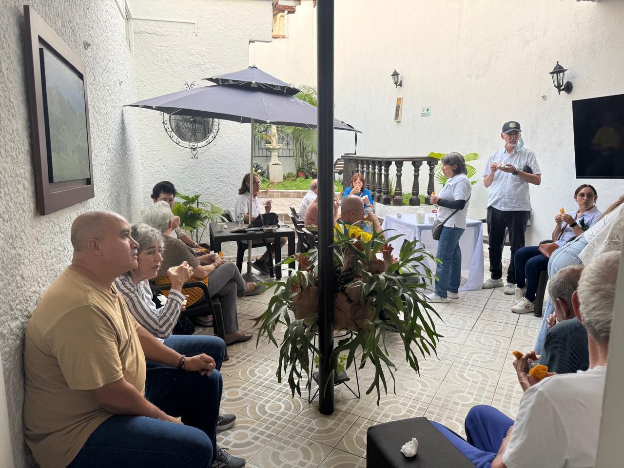 Grupo de personas en un patio al aire libre escuchando una presentación; hay plantas y una sombrilla.