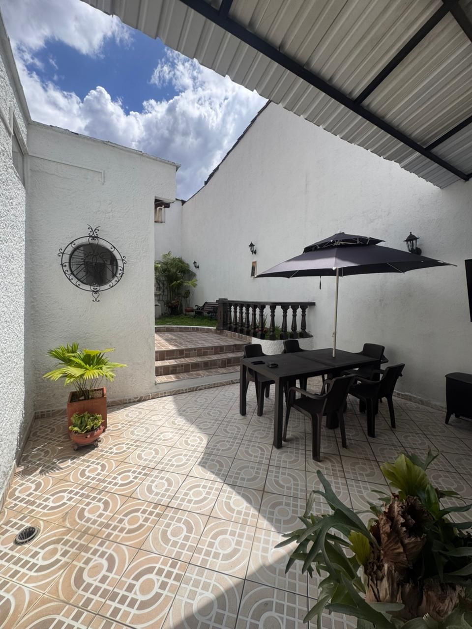 Patio con mesa, sillas, sombrilla y plantas en macetas contra paredes blancas; cielo azul visible arriba.