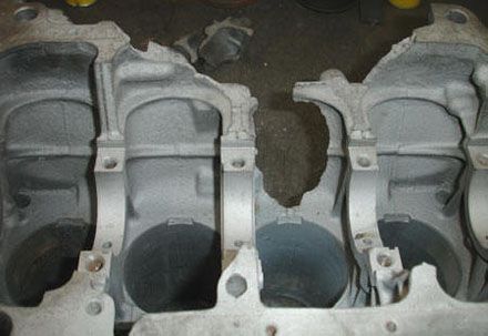 Bloque de motor dañado con fracturas visibles. Metal gris con agujeros cilíndricos.