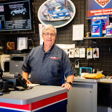 Man at auto shop counter, smiling, wearing mechanic shirt. Shop interior with car parts and signage | Rick's Automotive