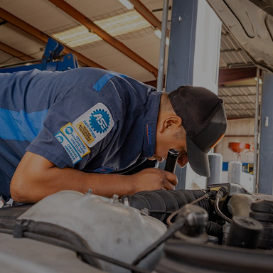 Mechanic in blue shirt and cap examines engine with a flashlight in a garage. | Rick's Automotive