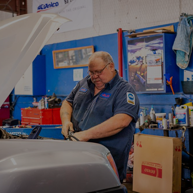 Mechanic working on a car in a shop, inspecting under the hood. Blue uniform, tools, and auto parts visible. | Rick's Automotive