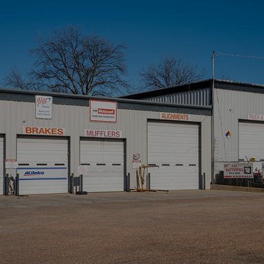 Auto repair shop with garage doors, signs for brakes, mufflers, and alignments. Clear blue sky. | Rick's Automotive
