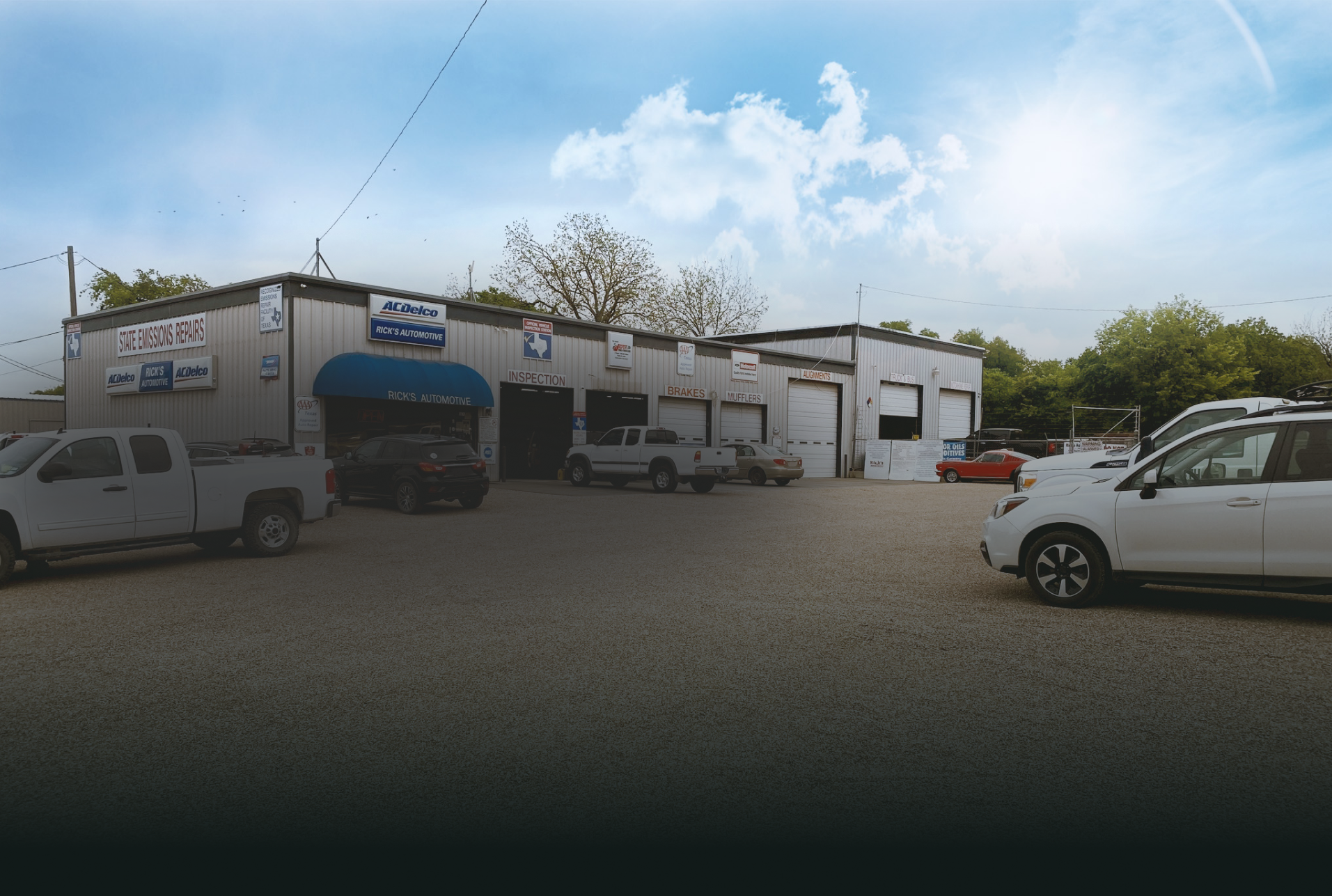 Auto repair shop exterior with cars in the lot under a bright blue sky. | Rick's Automotive