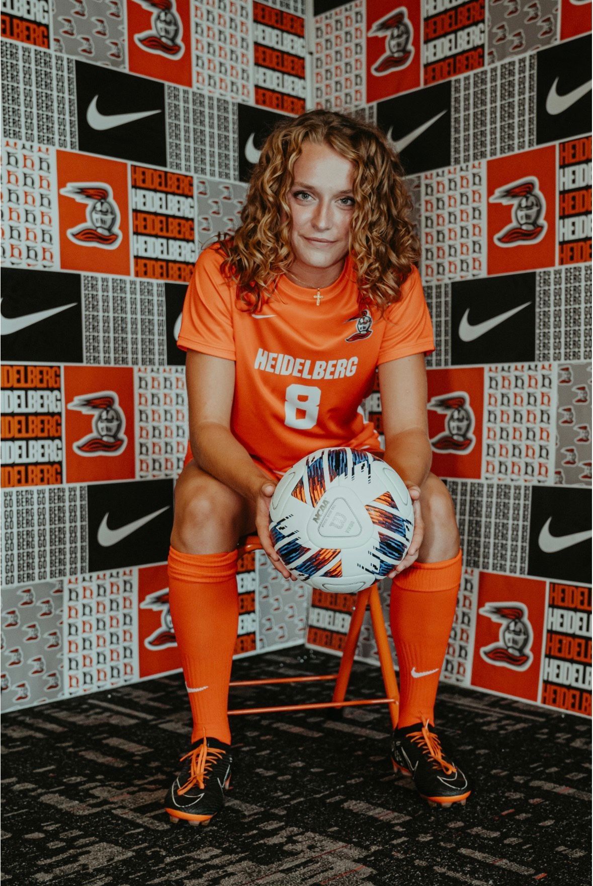 Soccer player in orange uniform holding a ball, sitting, with logo-covered wall.