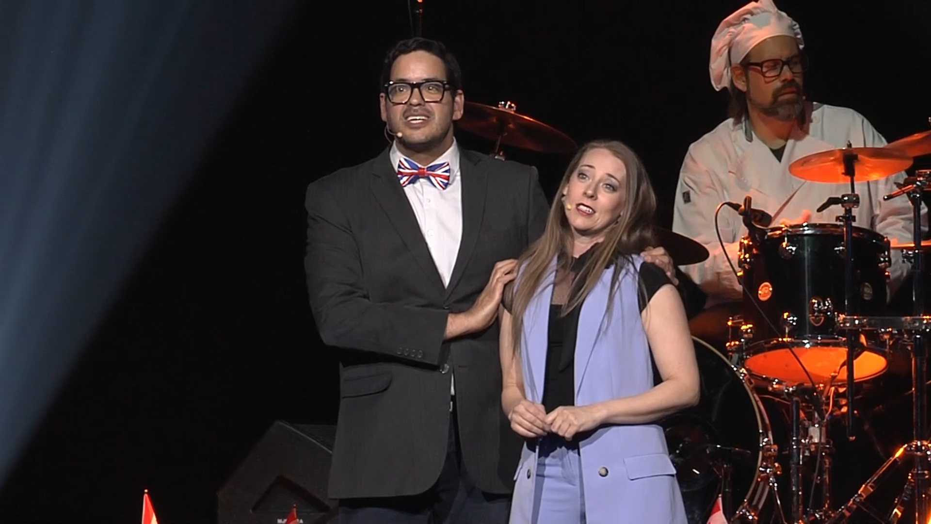 A man and a woman stand on a stage in front of a drum set