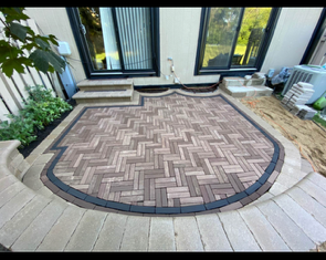 Brick paved area, gray bricks in herringbone pattern, bordered by light gray and dark gray pavers, near greenery.