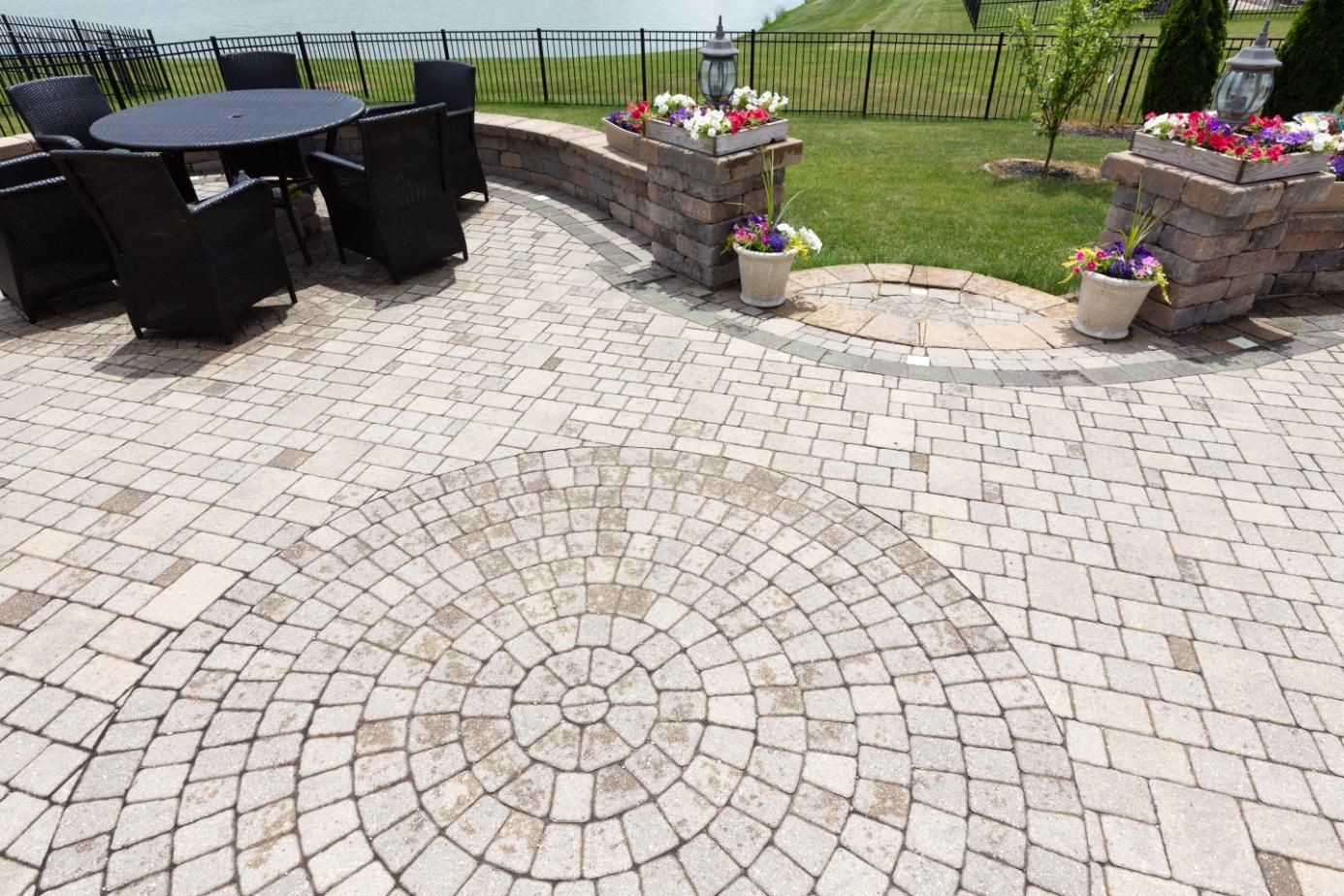 Circular brick patio with a round table and chairs, bordered by a low stone wall with flower pots and green lawn.