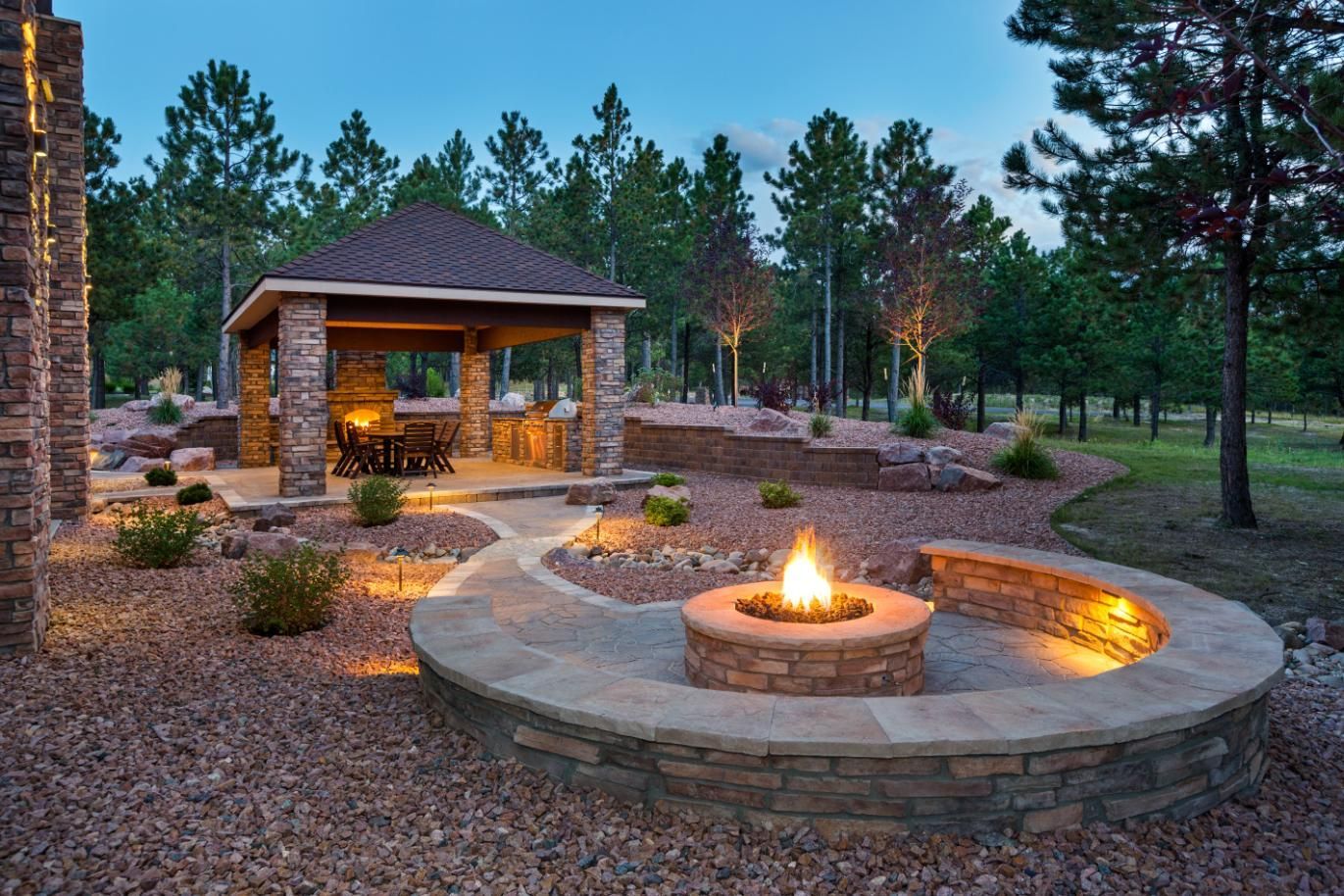 Stone fire pit and gazebo in a landscaped backyard at dusk, with trees in the background.