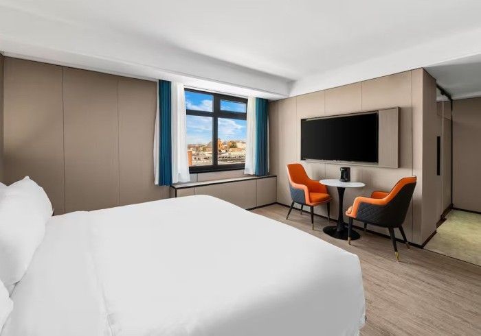 Hotel room with bed, chairs, table, large TV on wall, and a window with city view.