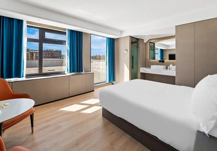 Hotel room with a bed, table, windows with blue curtains, and a bathroom visible.