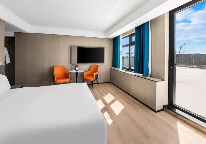 Hotel room with a bed, TV, orange chairs, and a window overlooking an outdoor area.