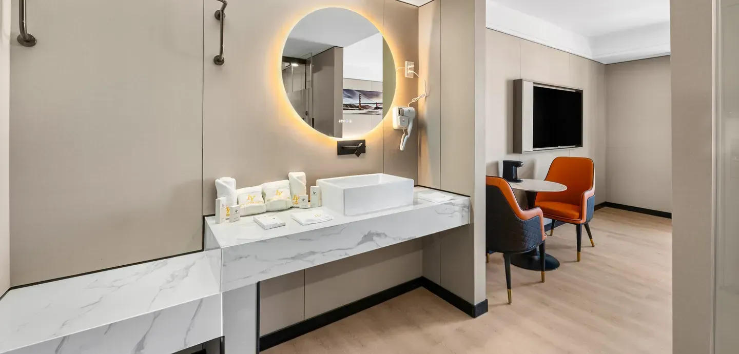 Modern hotel bathroom with marble countertop, round mirror, and seating area.