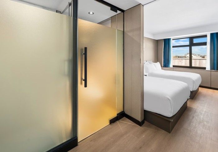 Hotel room with frosted glass shower door, two twin beds, and a window with blue curtains.