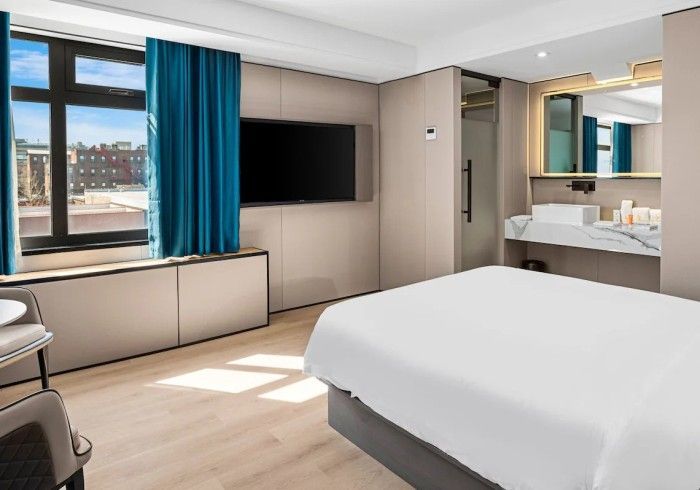 Modern hotel room with a bed, a large TV, and a window with teal curtains.