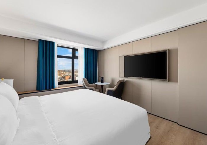 Hotel room with a bed, TV, and small table by a window with blue curtains and an outdoor view.