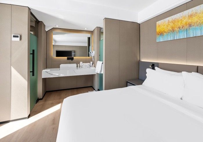 Hotel room with a bed, bathroom area, and painting on the wall. Bright natural light.