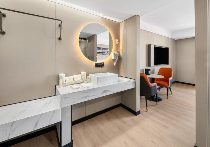 Modern hotel bathroom and sitting area with marble vanity, round mirror, and chairs.