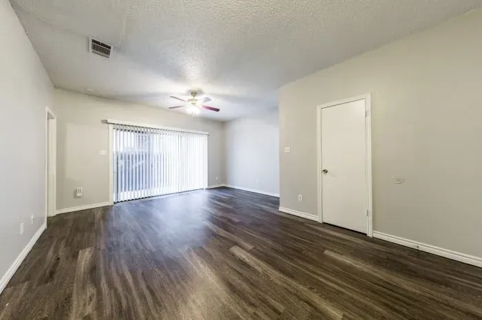 Three Villas - living room with grey laminate wood flooring, ceiling fan, and sliding door with blinds to back patio.